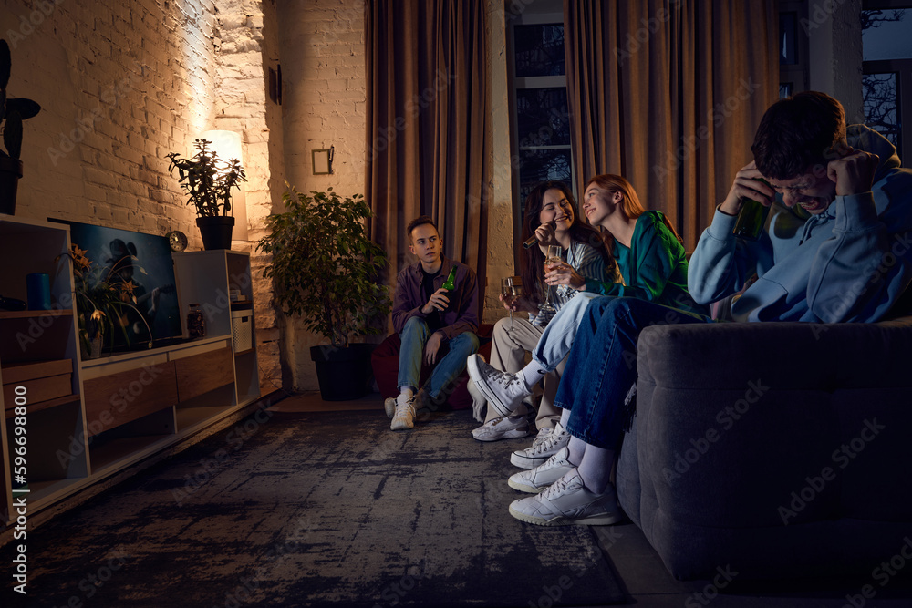 Group of friends, young people sitting on couch at home in the evening and singing karaoke, having fun, talking and drinking beer. Concept of friendship, leisure activity, weekends, fun, emotions