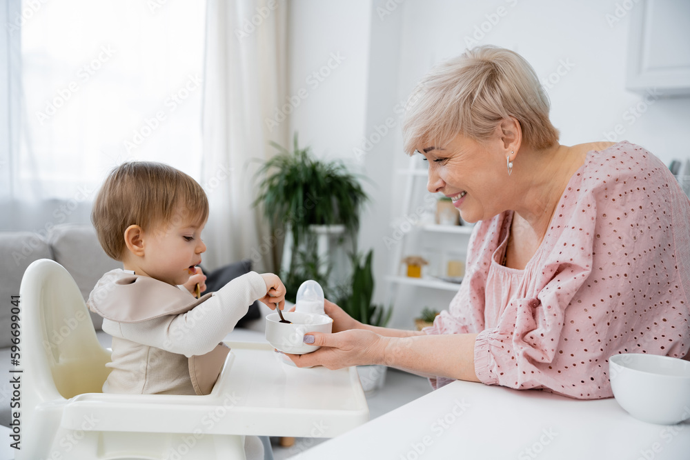 side view of smiling middle aged woman holding bowl near granddaughter sitting on baby chair and having breakfast in kitchen. side view of smiling middle aged woman holding bowl near granddaughter sitting on baby chair and having breakfast in kitchen.
