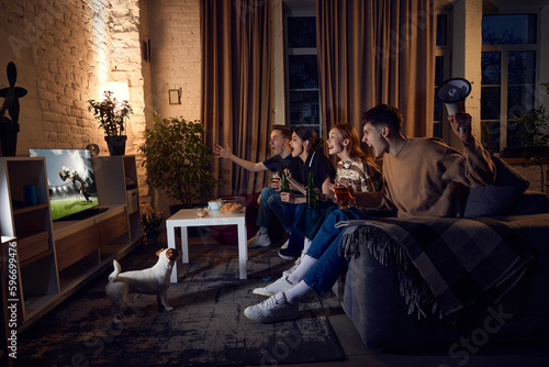 Group of friends, sport fans sitting on couch at home in the evening and emotionally watching online football match. Concept of friendship, leisure activity, weekends, fun, emotions