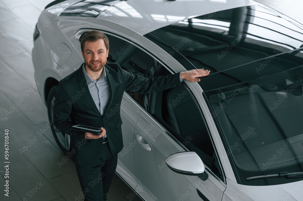 Top view, with tablet. Man in black suit is indoors in the car dealership near automobile Top view, with tablet. Man in black suit is indoors in the car dealership near automobile