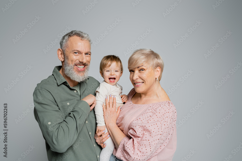 happy mature couple looking at camera near laughing granddaughter isolated on grey.