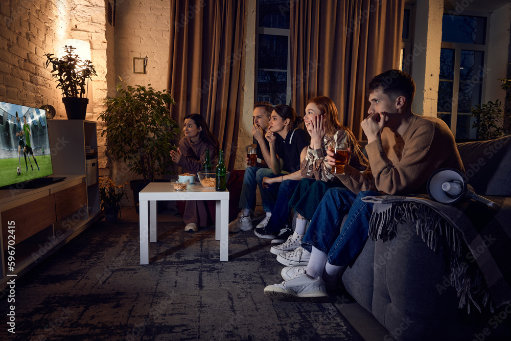 Group of young people, friends sitting on couch at home in the evening and watching live football match translation on tv. Concept of friendship, leisure activity, weekends, fun, emotions Group of young people, friends sitting on couch at home in the evening and watching live football match translation on tv. Concept of friendship, leisure activity, weekends, fun, emotions