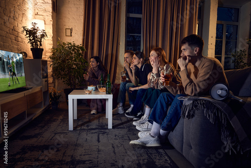 Group of young people, friends sitting on couch at home in the evening and watching live football match translation on tv. Concept of friendship, leisure activity, weekends, fun, emotions