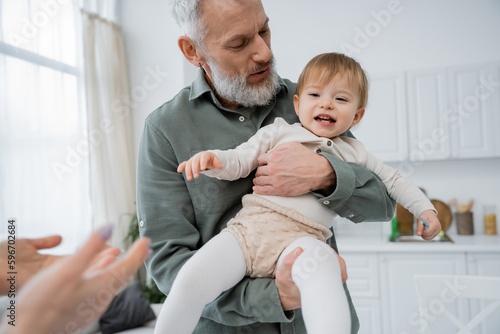 mature bearded man holding cheerful toddler granddaughter in kitchen.