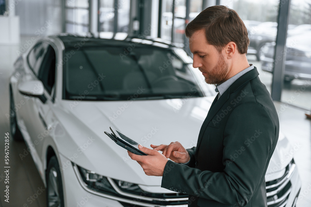 Standing, side view, holding tablet. Man in black suit is indoors in the car dealership near automobile Standing, side view, holding tablet. Man in black suit is indoors in the car dealership near automobile