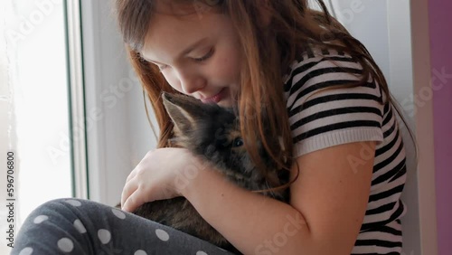 A little girl sitting on the windowsill touchingly embraces a brown rabbit, pressing her face to it, the love of children for pets.