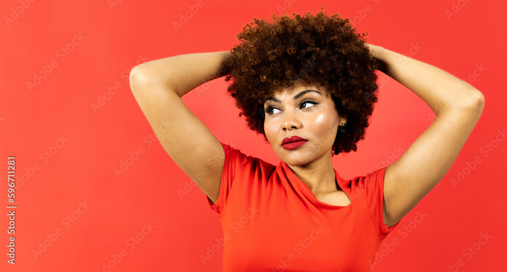 A dark-skinned girl with afro hair poses on a red background. The girl touches her hair with both hands while looking to the side. African fashion concept for girls with afro hair. A dark-skinned girl with afro hair poses on a red background. The girl touches her hair with both hands while looking to the side. African fashion concept for girls with afro hair.