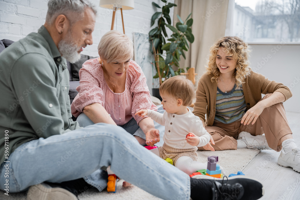 joyful woman looking at middle aged parents playing building blocks game with child on floor in living room. joyful woman looking at middle aged parents playing building blocks game with child on floor in living room.