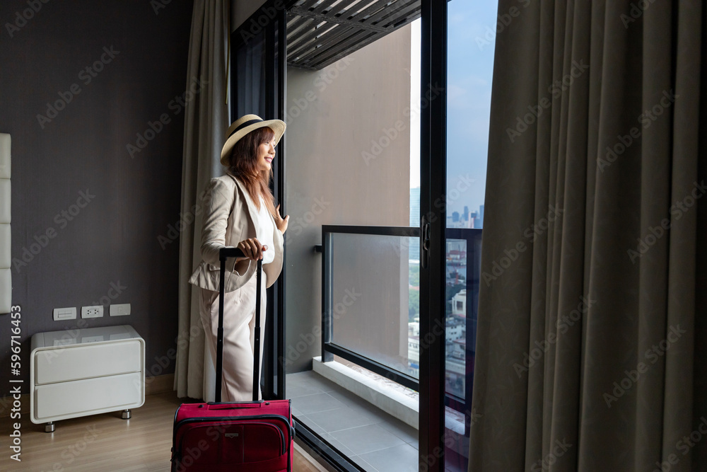 Asian woman moving the luxury hotel with luggage on her vacation while looking at the view from window with high rise building in the urban downtown area for business trip and condominium living usage