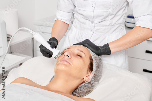 Fotografie Cosmetologist makes ultrasound skin tightening for rejuvenation woman face using phonophoresis, anti aging cosmetic procedure with in beauty spa salon