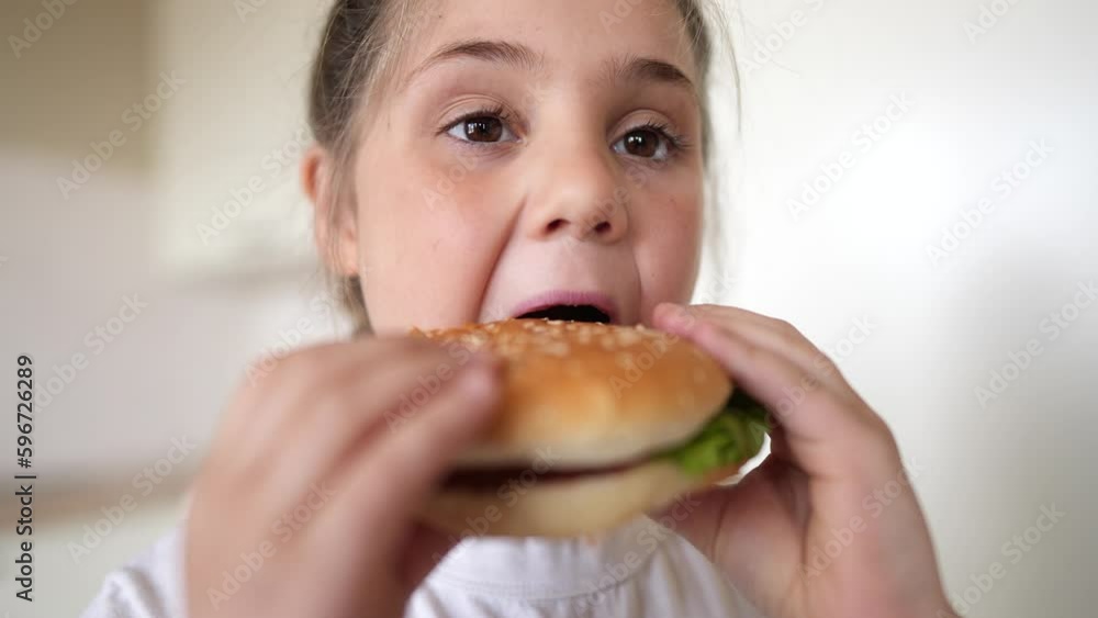 little girl eating a hamburger. unhealthy fast food proper nutrition concept. child greedily with pleasure bites a big burger in the kitchen meal at home. kid eats fast food close-up little girl eating a hamburger. unhealthy fast food proper nutrition concept. child greedily with pleasure bites a big burger in the kitchen meal at home. kid eats fast food close-up