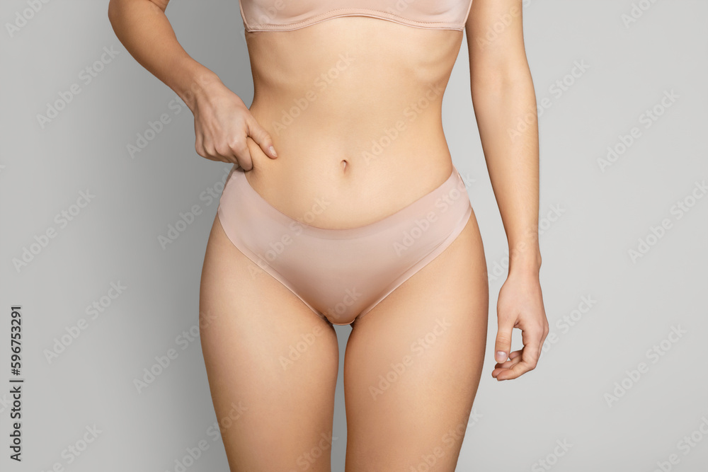 Unrecognizable lady in beige underwear showing excessive fat on her belly, woman pulling skin on her waist, cropped