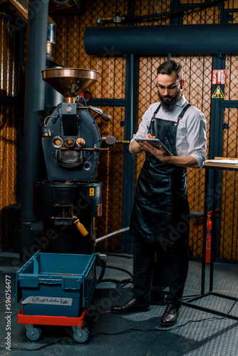 serious guy in apron standing in coffee production near machine for roasting coffee beans with tablet in hands