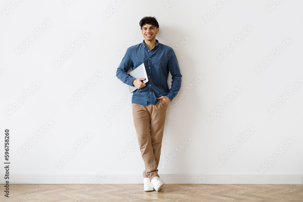 Cheerful Middle Eastern Man Holding Laptop Standing Near Wall Indoors Cheerful Middle Eastern Man Holding Laptop Standing Near Wall Indoors