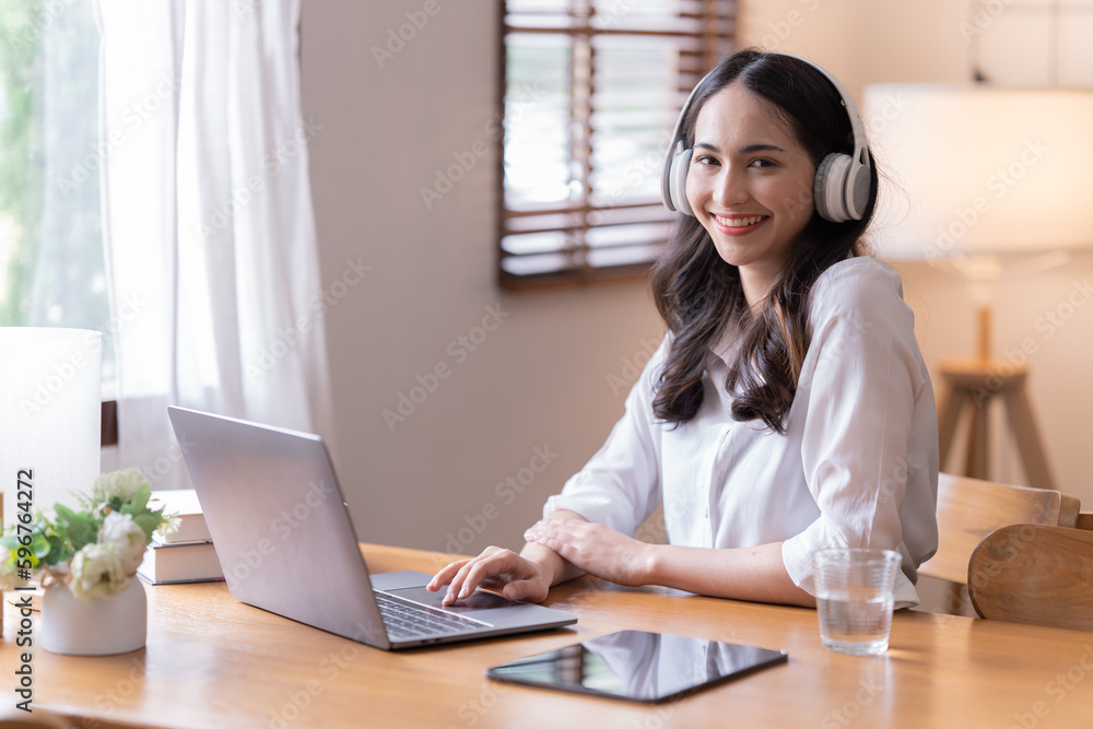 Attractive happy young Asian businesswomen at home, sitting at the desk, using a laptop computer, tablet and headphones having a video chat. Attractive happy young Asian businesswomen at home, sitting at the desk, using a laptop computer, tablet and headphones having a video chat.