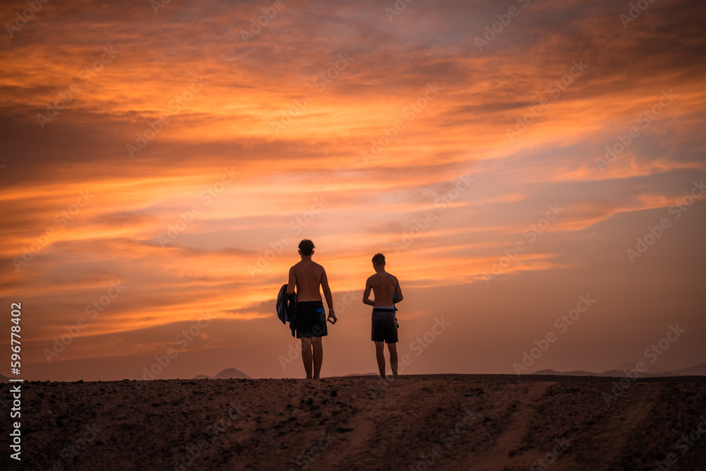 Nähe Marsa Alam, Ägypten - Herbst - 2 Jugendliche in der Wüste in Badeshorts, Sonnenuntergang und die Red Sea Mountains im Hintergrund.