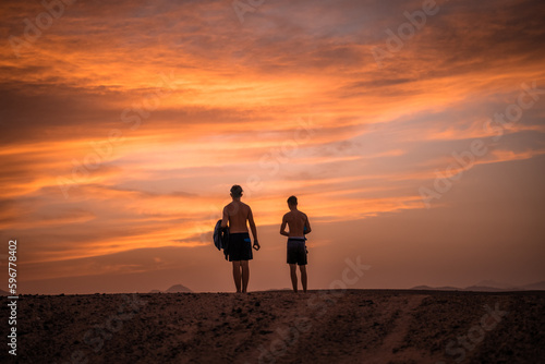 Nähe Marsa Alam, Ägypten - Herbst - 2 Jugendliche in der Wüste in Badeshorts, Sonnenuntergang und die Red Sea Mountains im Hintergrund.