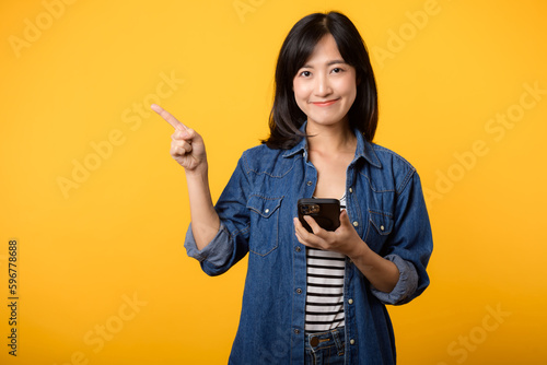 Obraz na plátně Portrait beautiful young asian woman happy smile dressed in denim jacket showing smartphone with pointing finger hand gesture to free space isolated on yellow studio background