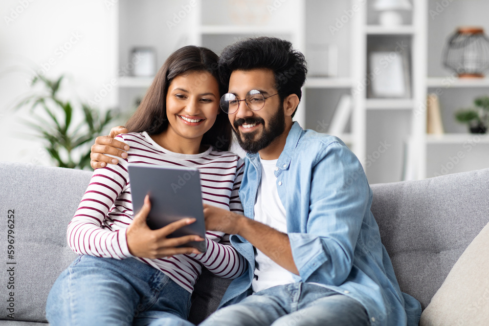 Happy loving indian couple sitting on couch, using digital pad Happy loving indian couple sitting on couch, using digital pad