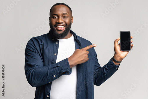 Excited Africal man with smartphone in the hand isolated on gray background, pointing finger on phone with empty screen, advertising mobile app, showing deal photo