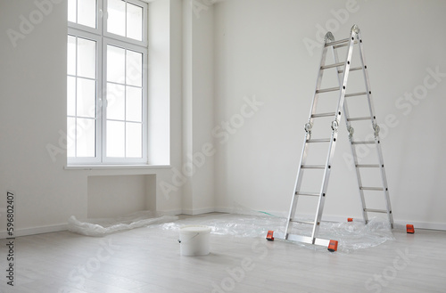 Modern home after repairs and renovations. New empty house interior with white freshly painted walls, step ladder, paint can, protective plastic on the floor, big window, and no people