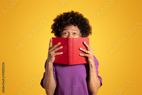 Cheerful excited adult african american curly man in purple t-shirt looks from behind book