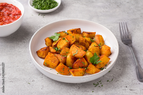 Traditional Spanish potato, patatas bravas with smoked paprika, spicy tomato sauce and parsley in a white bowl on a gray stone background, top view. Delicious homemade food