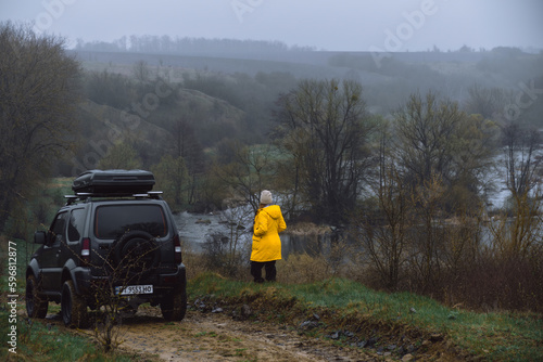 Khmelnytskyi, Ukraine - April 15, 2023: Suzuki Jimny off-road vehicle, dark gray, roof rack Niken 390, spring day, landscape with river. A car for extreme adventures. Women in yellow jacket
