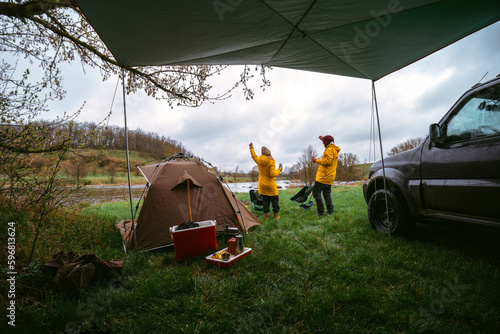Happy couple, clink beer cans. in wilderness. The celebrating good trip, the tent and tourist equipment. Yellow raincoats from bad weather. A gloomy spring day. Camping by off road vehicle. River