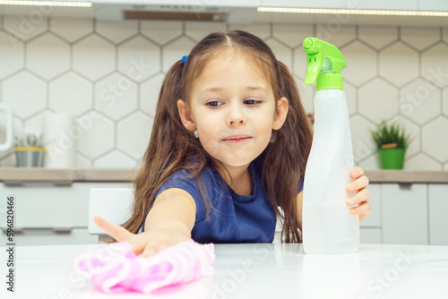 Portrait of focused little preteen girl with dark hair wearing blue T-shirt sanitizing polishing white table with rag near spray detergent in kitchen. Housework, housekeeping, sanitizing, hygiene.