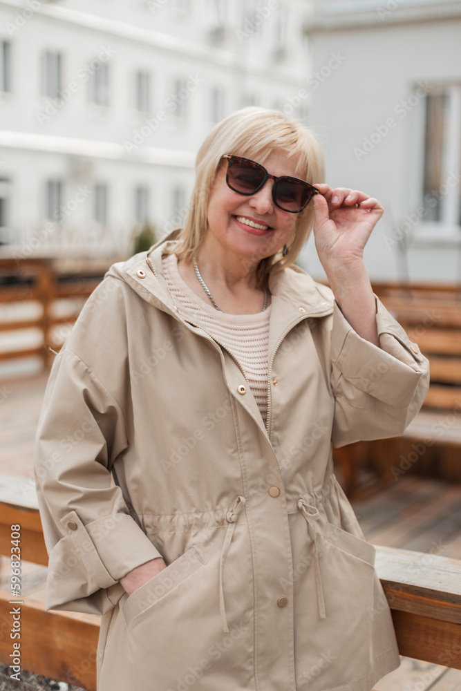 Happy beautiful senior woman with a smile in fashionable casual beige clothes with a jacket puts on sunglasses and walks in the city