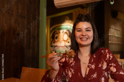Obraz na plátně She is dressed in Japanese print clothes and holds an octopus ball with her chopsticks while smiling