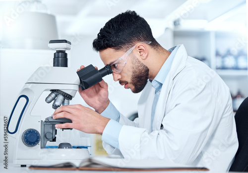 One serious young male medical scientist sitting at a desk and using a microscope to examine and analyse test samples on slides in a hospital. Hispanic healthcare biochemist professional discovering