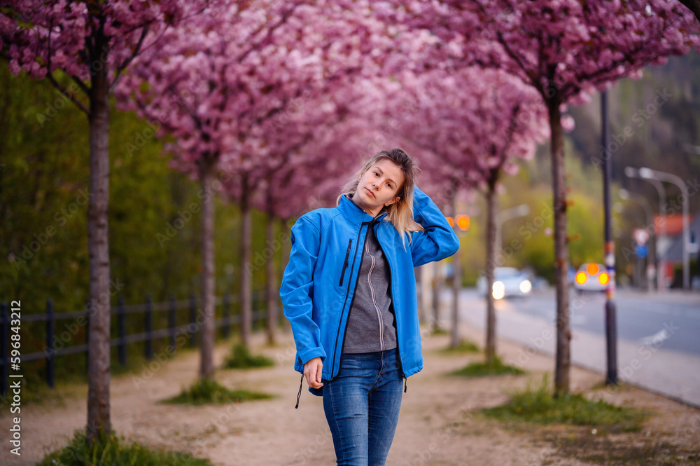 Fototapeta premium Yong blonde woman in blue jacket and jeans stands in pink blooming sacura alley