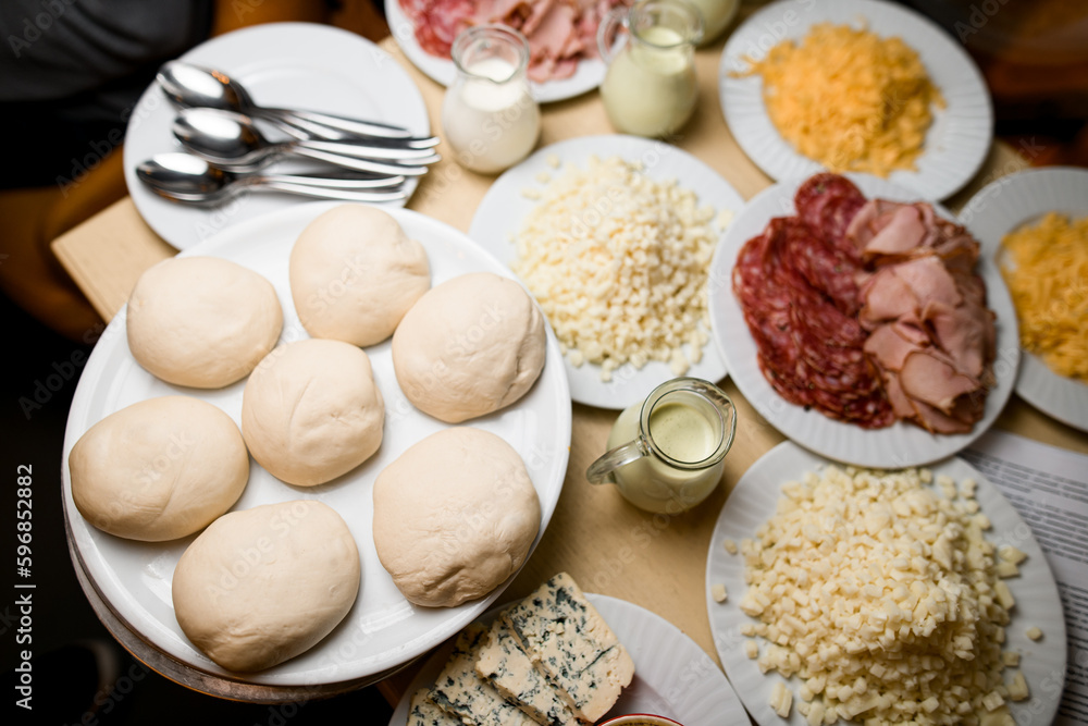 Uncooked mini pizzas and ingridients for them - salami, jamon, sause, cheese on plates. Front view