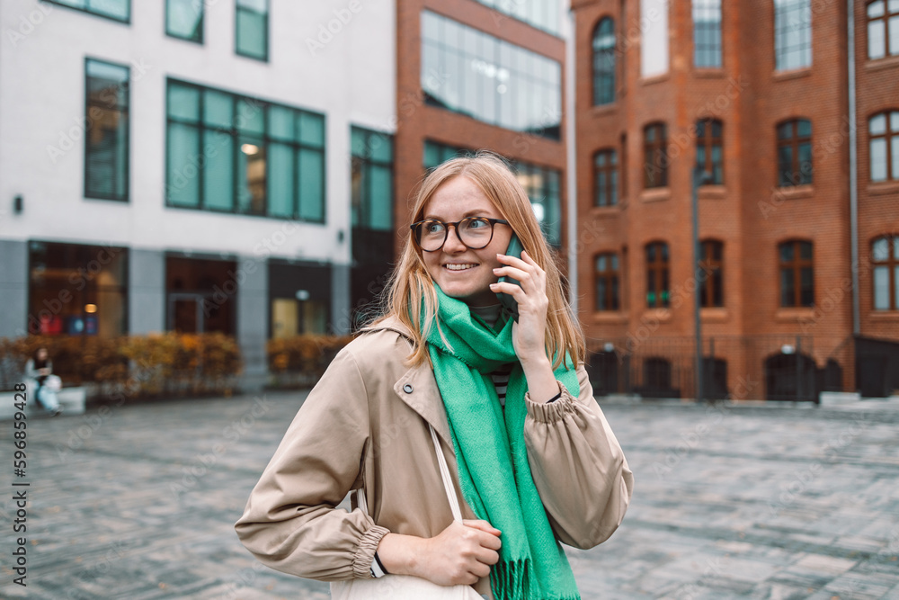 Fototapeta premium Beautiful young woman talking on smartphone walking through city street. Portrait of gorgeous smiling female using mobile phone. High quality photo
