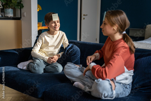 Fotografi Two kids brother and sister playing guessing game Who am I, sitting on sofa in living room with paper stickers on forehead