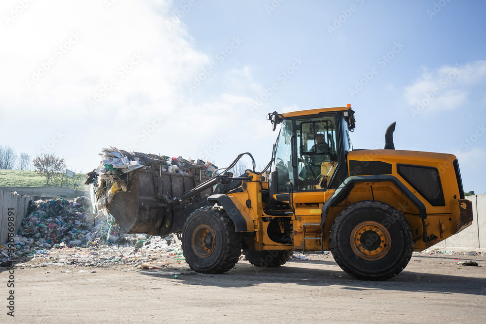 Yellow skid steer loader moving wooden waste material, shaking out a scrap grapple on the garbage heap in the materials recovery facility