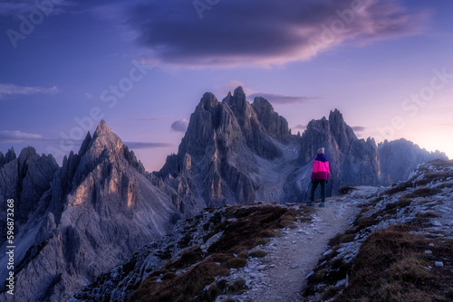 Woman on the hill and mountain peaks at sunset in spring in Dolomites, Italy. Girl on the trail and high rocks at dusk in fall. Colorful landscape with cliffs, stones, purple sky. Trekking and hiking