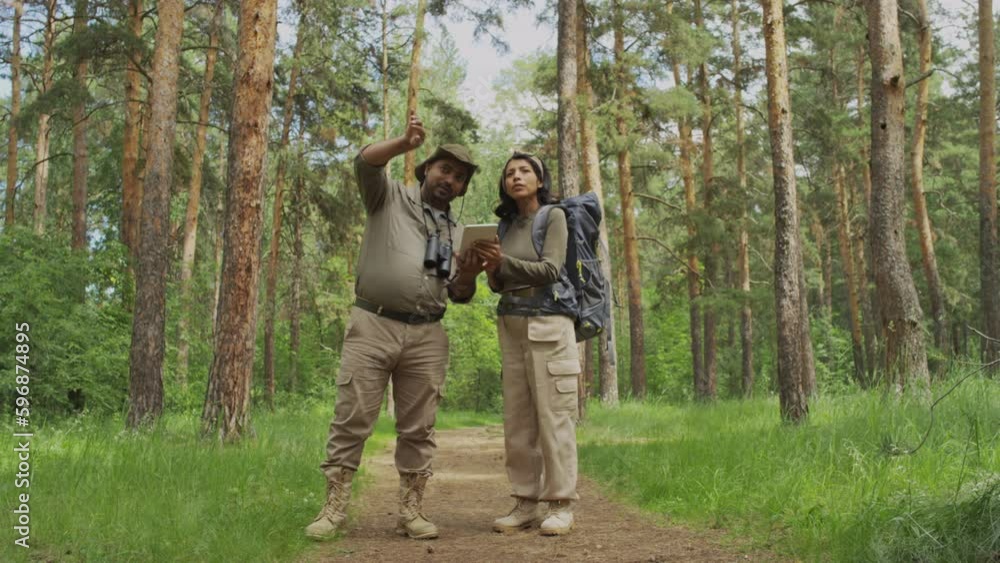 Full shot of multiracial male forest ranger in khaki uniform giving directions and pointing right way to female hiker with backpack and tablet computer, then woman thanking him and walking away Full shot of multiracial male forest ranger in khaki uniform giving directions and pointing right way to female hiker with backpack and tablet computer, then woman thanking him and walking away