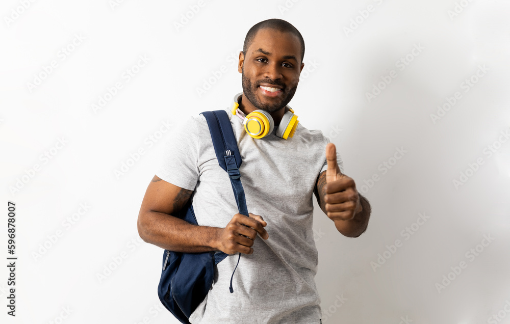 A dark-skinned man poses in casual clothing against a white background with headphones and a backpack. Man shows hand with thumb up, apt, happy with exam marks. A dark-skinned man poses in casual clothing against a white background with headphones and a backpack. Man shows hand with thumb up, apt, happy with exam marks.
