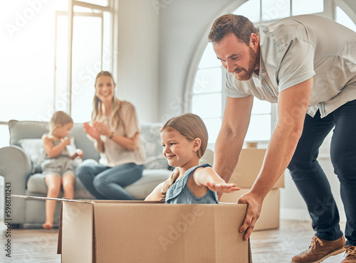 A happy mature caucasian father pushing his daughter in a box while her mother and sister sit on the sofa in the lounge at home. Man and girl having fun, playing games at home while enjoying family