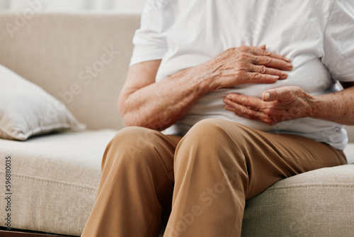 Elderly woman severe chest pain sitting on the sofa  health problems in old age  poor quality of life. Grandmother with gray hair holds her chest with her hands  women s health  breast cancer.