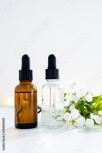 Two clear and amber glass cosmetic dropper bottles and white cherry flowers on white table  care products mockup