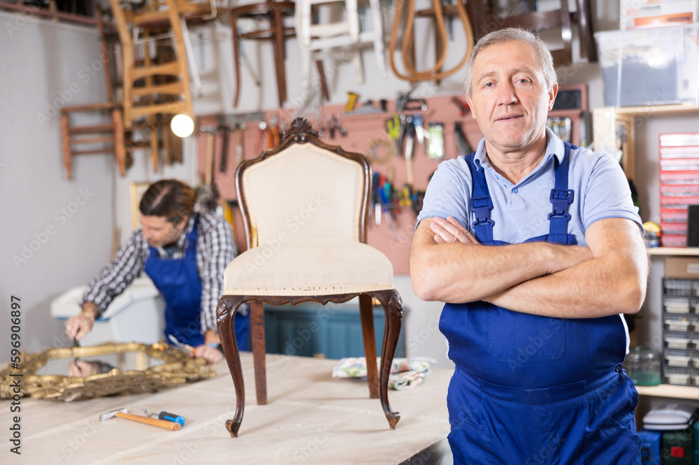Confident successful craftsman restorer in uniform looking at camera with crossed hands in furniture repair shop