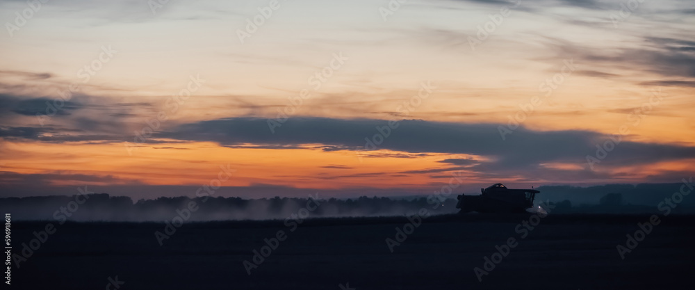 Silhouette of harvester machine to harvest wheat on sunset. Combine harvester driving on field on sunrise. Beautiful dawn sky above wheat field. Combine working in dusk. Wonderful twilight landscape.