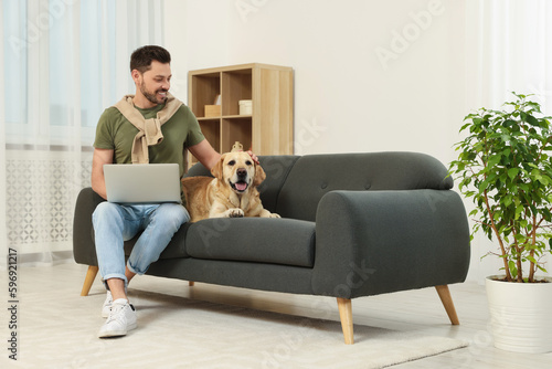 Man using laptop on sofa near his cute Labrador Retriever at home. Space for text