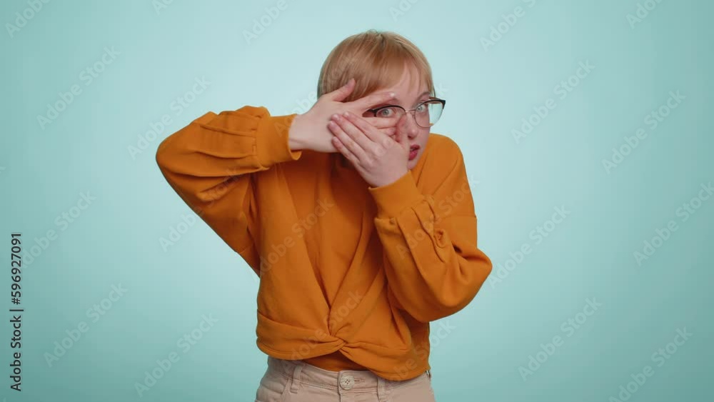 Nosy curious young woman in glasses closing eyes with hand and spying through fingers, hiding and peeping, binocular gesture exploring way seeking something in distance. Blonde girl on blue background Nosy curious young woman in glasses closing eyes with hand and spying through fingers, hiding and peeping, binocular gesture exploring way seeking something in distance. Blonde girl on blue background