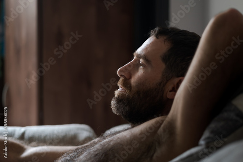 Leaning back shirtless on the sofa, a handsome man expresses being totally relaxed during a casual day at home. His thick beard, chest hair, and underarm hair are part of his look.