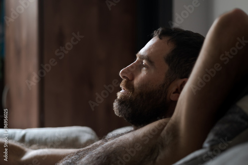 Leaning back shirtless on the sofa, a handsome man expresses being totally relaxed during a casual day at home. His thick beard, chest hair, and underarm hair are part of his look. photo